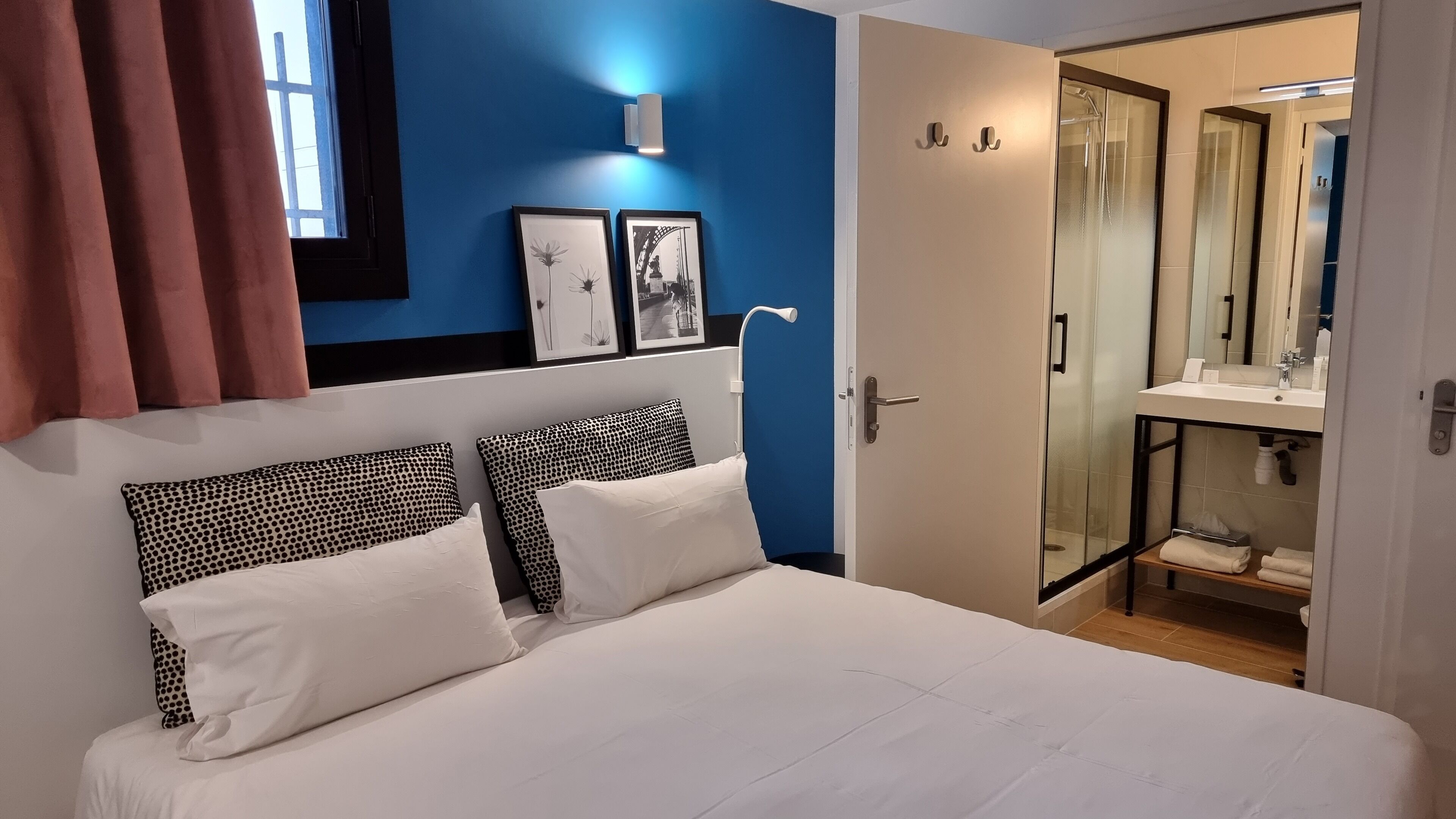 Un dormitorio con cama, dos almohadas, una mesita de noche, una ventana con cortinas, una lámpara fijada a la pared y una puerta que da a un baño con cabina de ducha de vidrio.