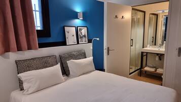 Un dormitorio con cama, dos almohadas, una mesita de noche, una ventana con cortinas, una lámpara fijada a la pared y una puerta que da a un baño con cabina de ducha de vidrio.