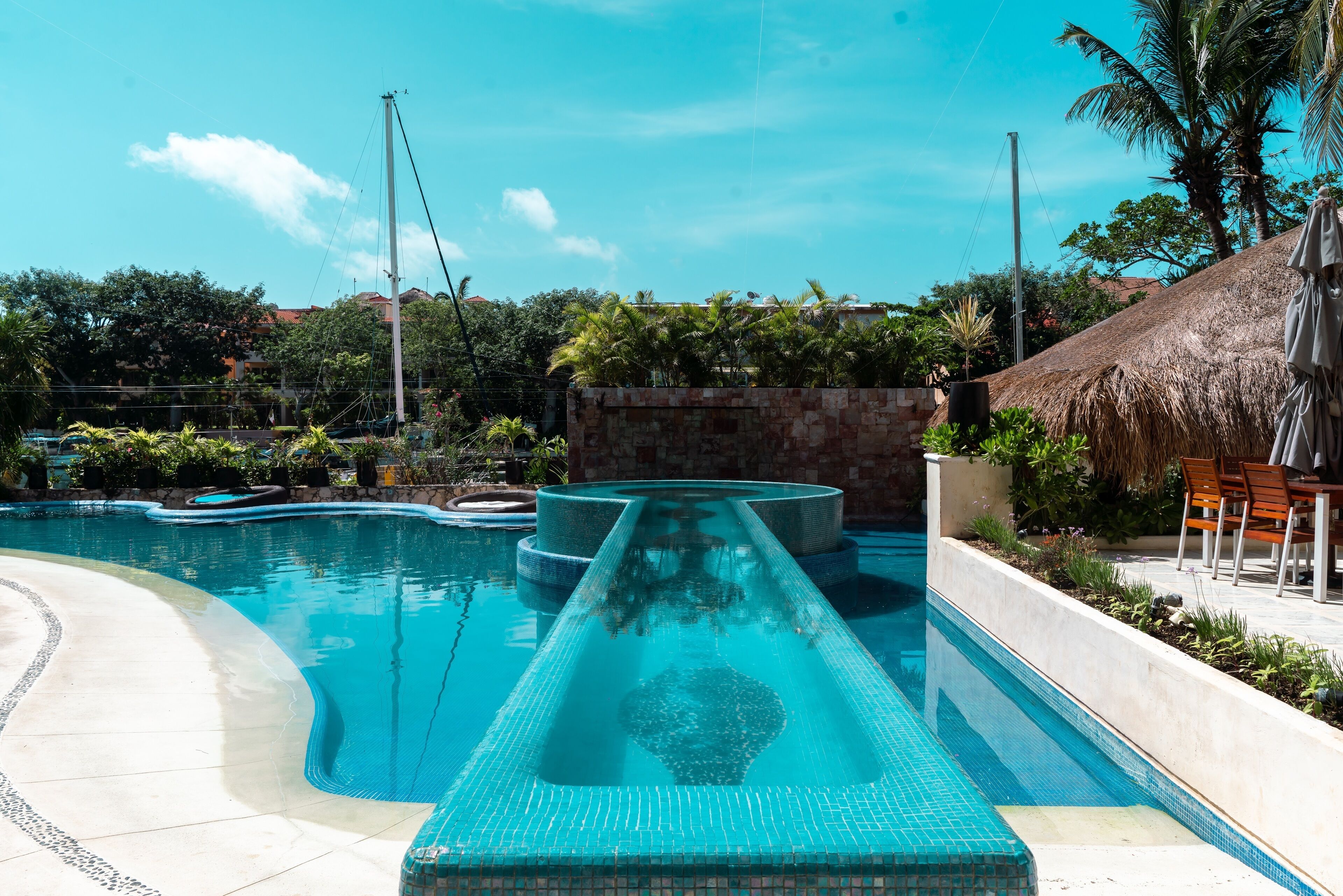 A swimming pool with a water slide, surrounded by palm trees and a thatched-roof structure.