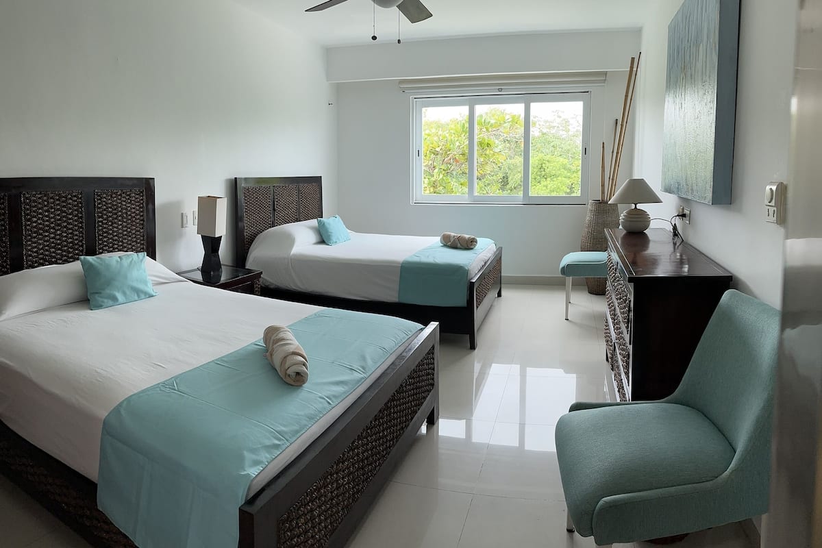 Une chambre d’hôtel avec deux lits, un ventilateur de plafond, une grande fenêtre et un bureau avec une lampe.