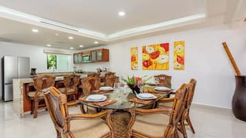 Un comedor moderno con una mesa redonda de vidrio rodeada de sillas de mimbre, una cocina al fondo y tres cuadros con motivos florales en la pared.