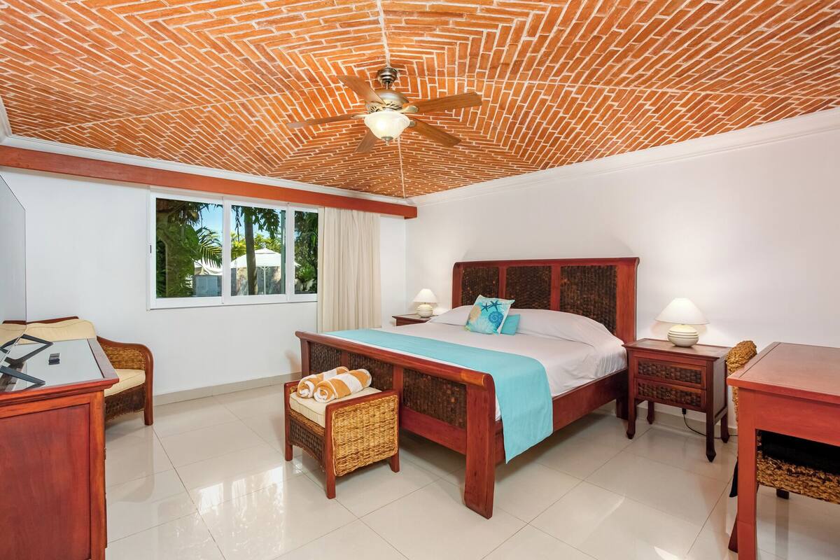 Une chambre d’hôtel avec un plafond orné d’un motif en briques, un lit avec une tête de lit en bois, un fauteuil en osier, un bureau et une fenêtre donnant sur un paysage verdoyant.