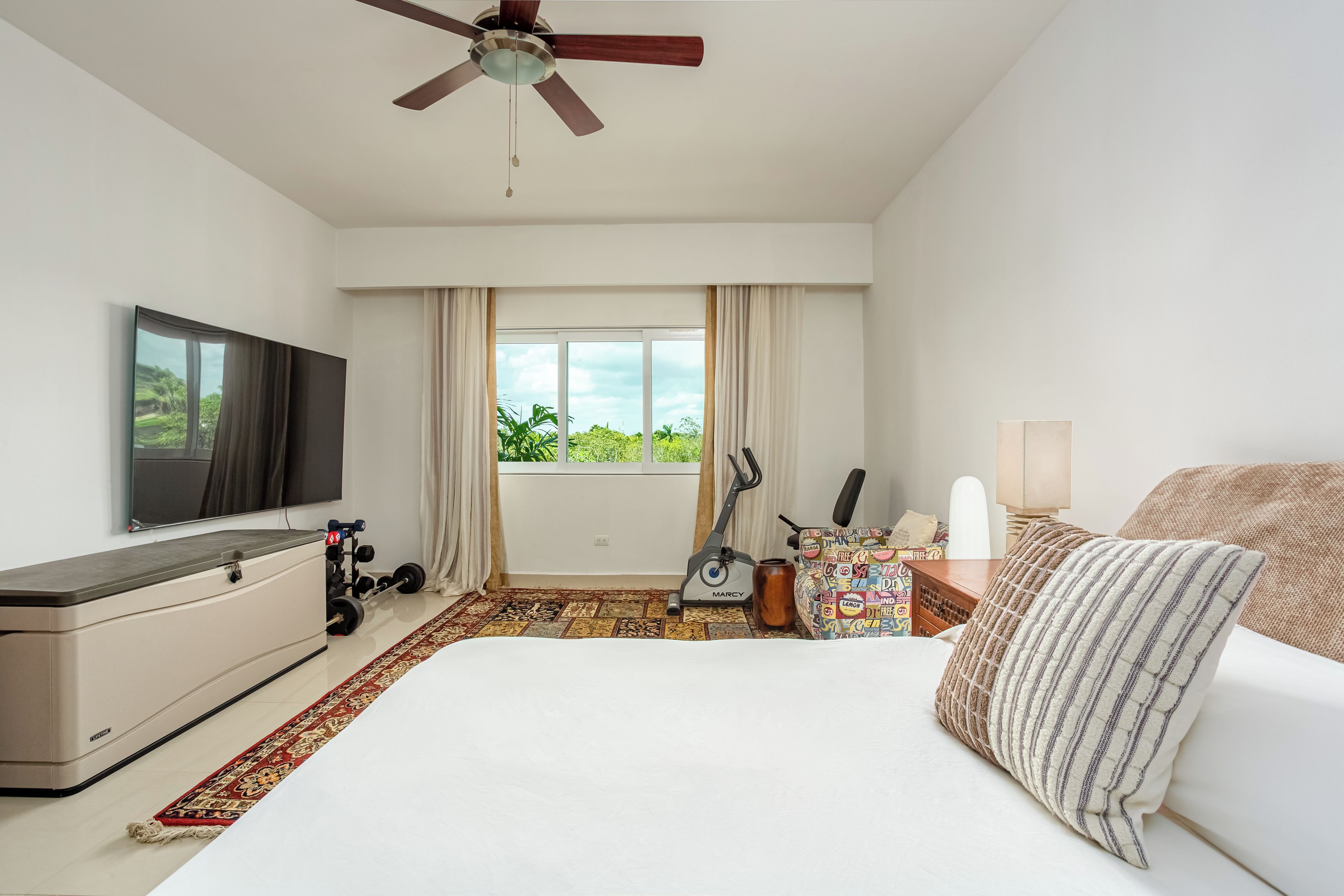 A hotel room with a bed, a TV, a ceiling fan, a window with curtains, a workout station, and a view of greenery outside.