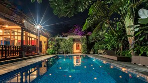 A nighttime swimming pool with a tiled deck, surrounded by lush greenery and a traditional building with a tiled roof. - Villa Mulyono (Batu)