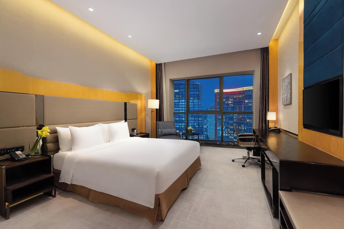 Une chambre d’hôtel avec un grand lit, un bureau avec une chaise, une télévision et une vue sur le paysage urbain la nuit.