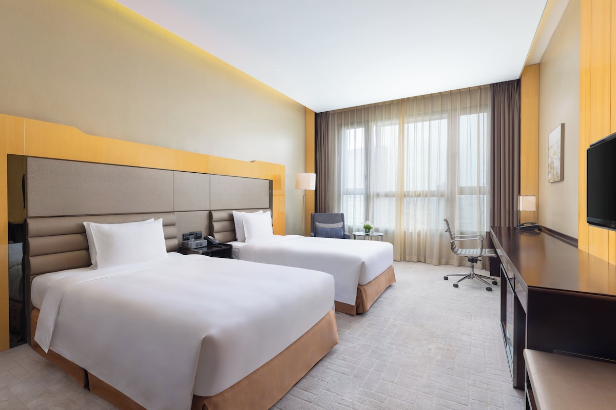 Quarto de hotel com duas camas, uma escrivaninha, uma cadeira, televisão e uma janela grande com cortinas.