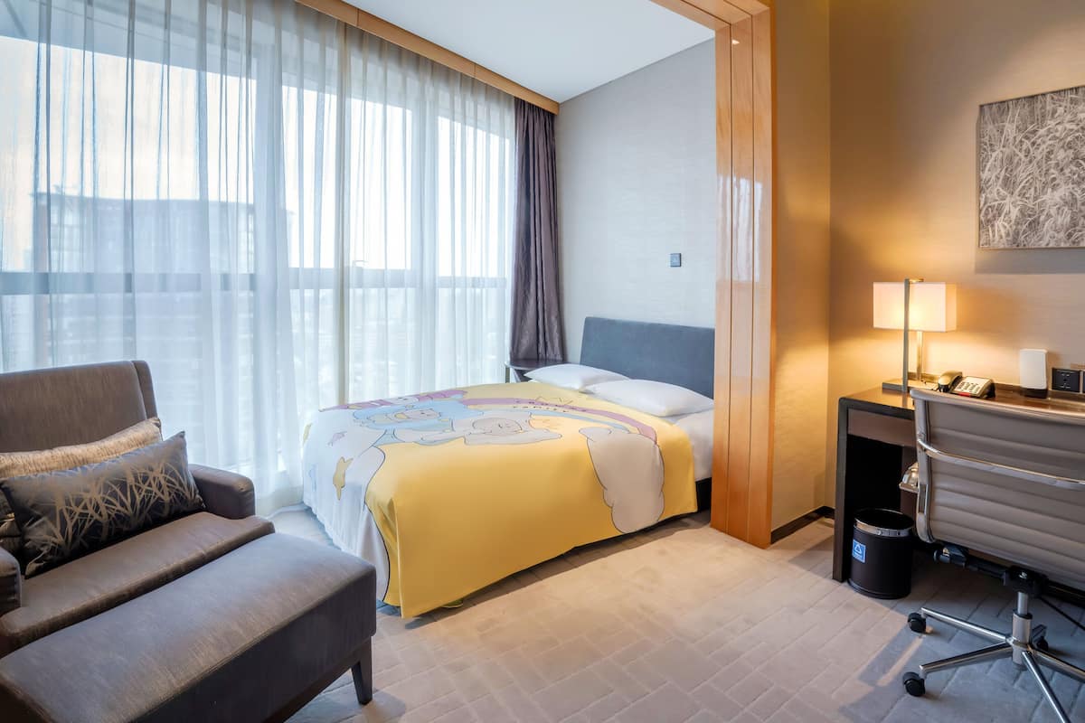Une chambre d’hôtel avec un lit, un bureau, une chaise et une fenêtre avec des rideaux légers.