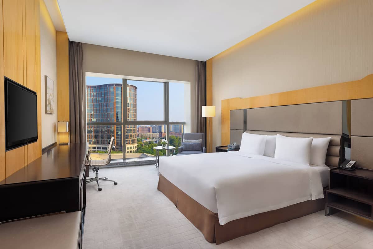 Une chambre d’hôtel avec un grand lit, un bureau, une chaise, une télévision et une vue sur le paysage urbain grâce à de grandes fenêtres.