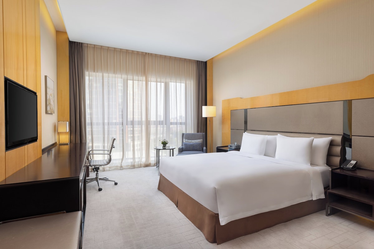 Quarto de hotel com uma cama grande, uma escrivaninha, uma cadeira, uma televisão, uma luminária e uma janela com cortinas.