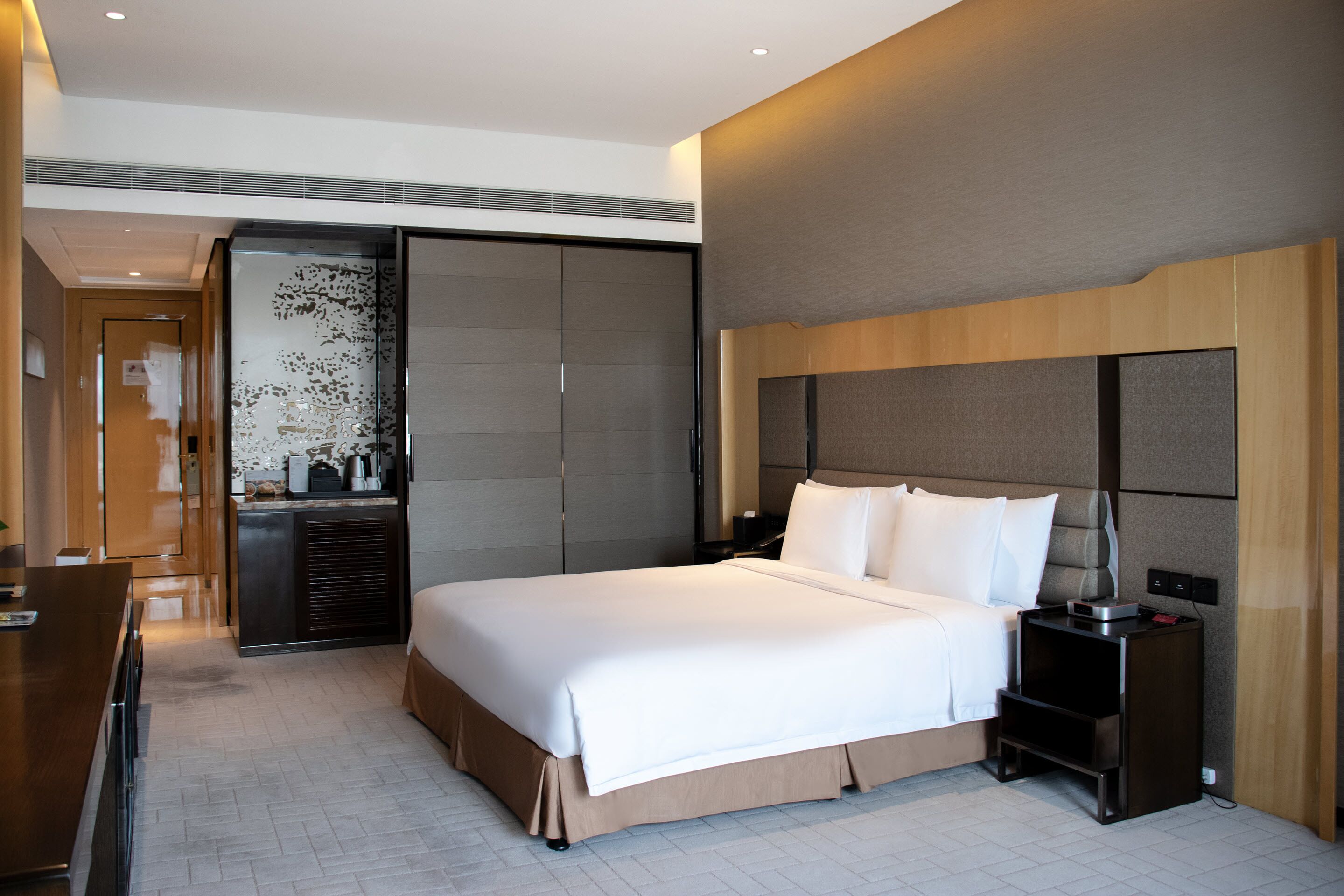 A modern hotel room with a large bed, a desk, a wardrobe, and a small table with a lamp.
