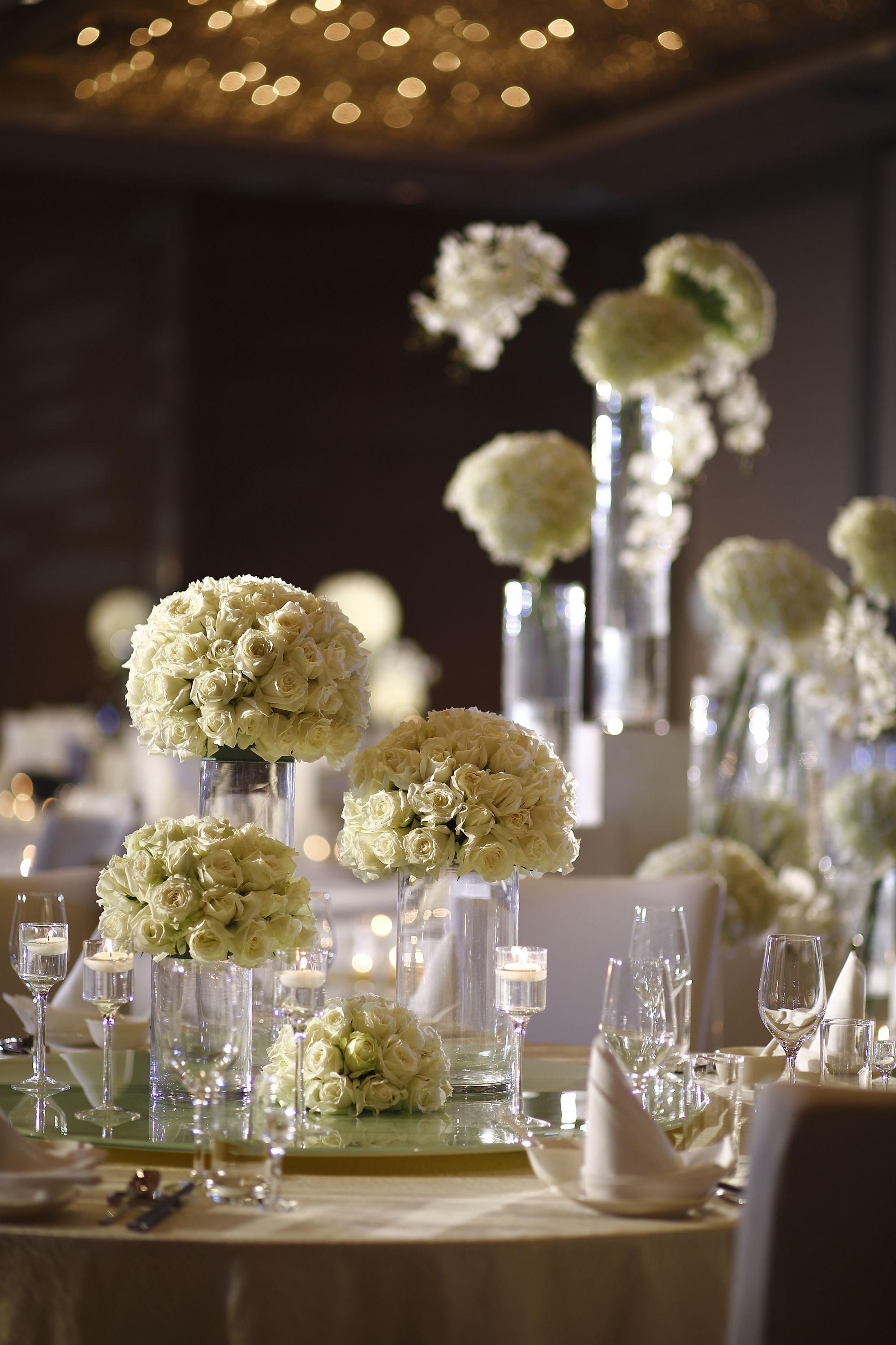 Una mesa adornada con flores blancas en altas jaras de vidrio, cristalería transparente y velas.