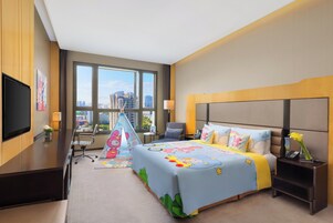 Habitación de hotel con una cama grande, un escritorio con televisor de pantalla plana, una silla, una mesita de noche con teléfono y una ventana con vista a la ciudad.
