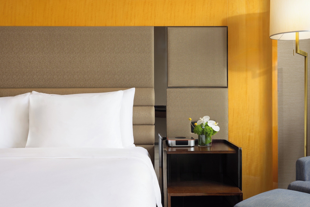 Quarto de hotel com cama, mesa de cabeceira, abajur e uma pequena mesa com um vaso de flores.