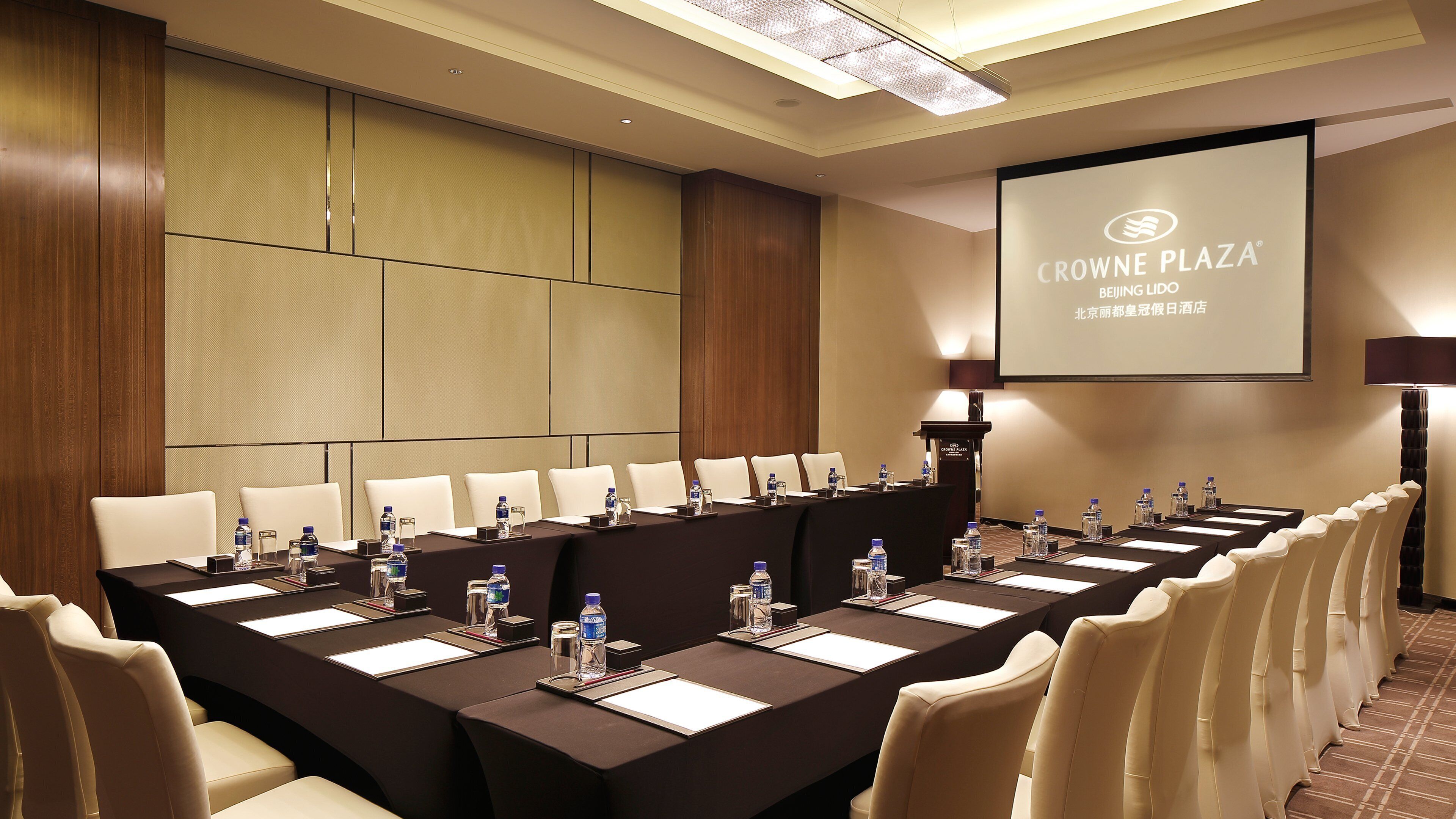 Un salón de conferencias con una larga mesa, múltiples sillas, botellas de agua y una pantalla proyectando "Crowne Plaza Beijing Lido".