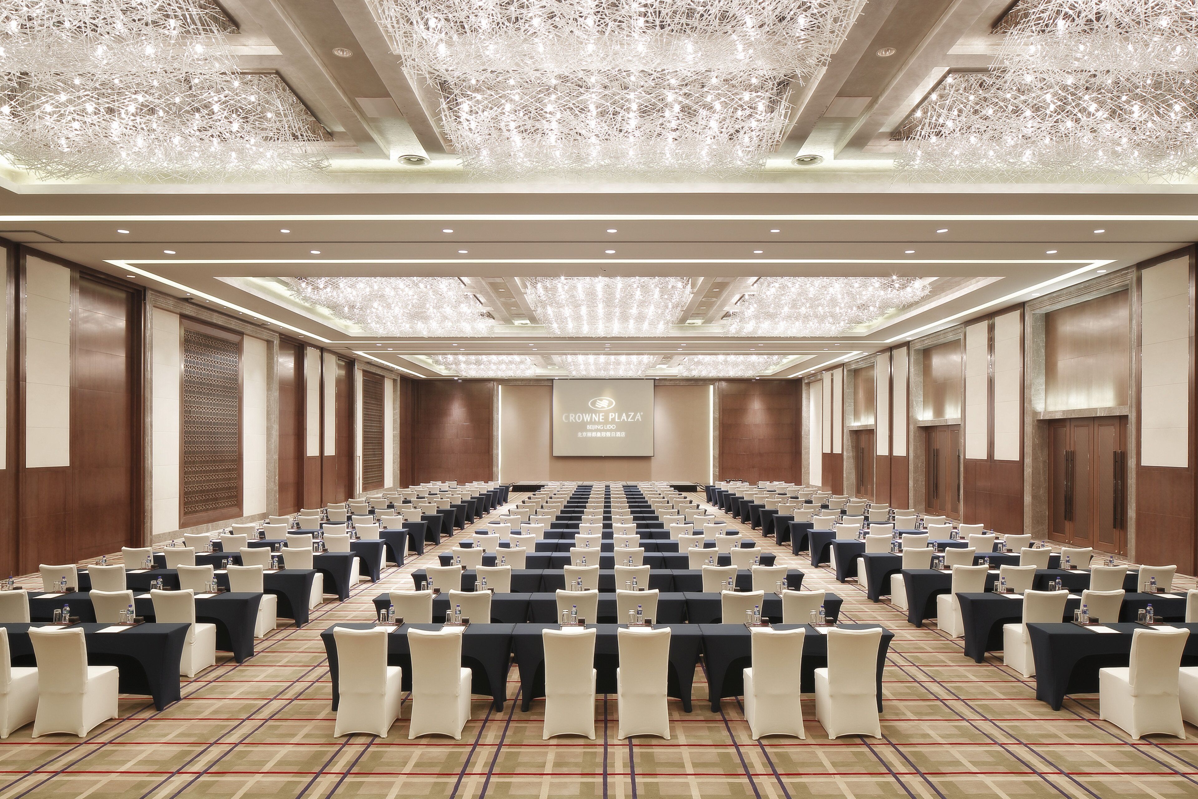 A conference room with rows of tables and chairs, a large screen at the front, and a ceiling with intricate lighting fixtures.