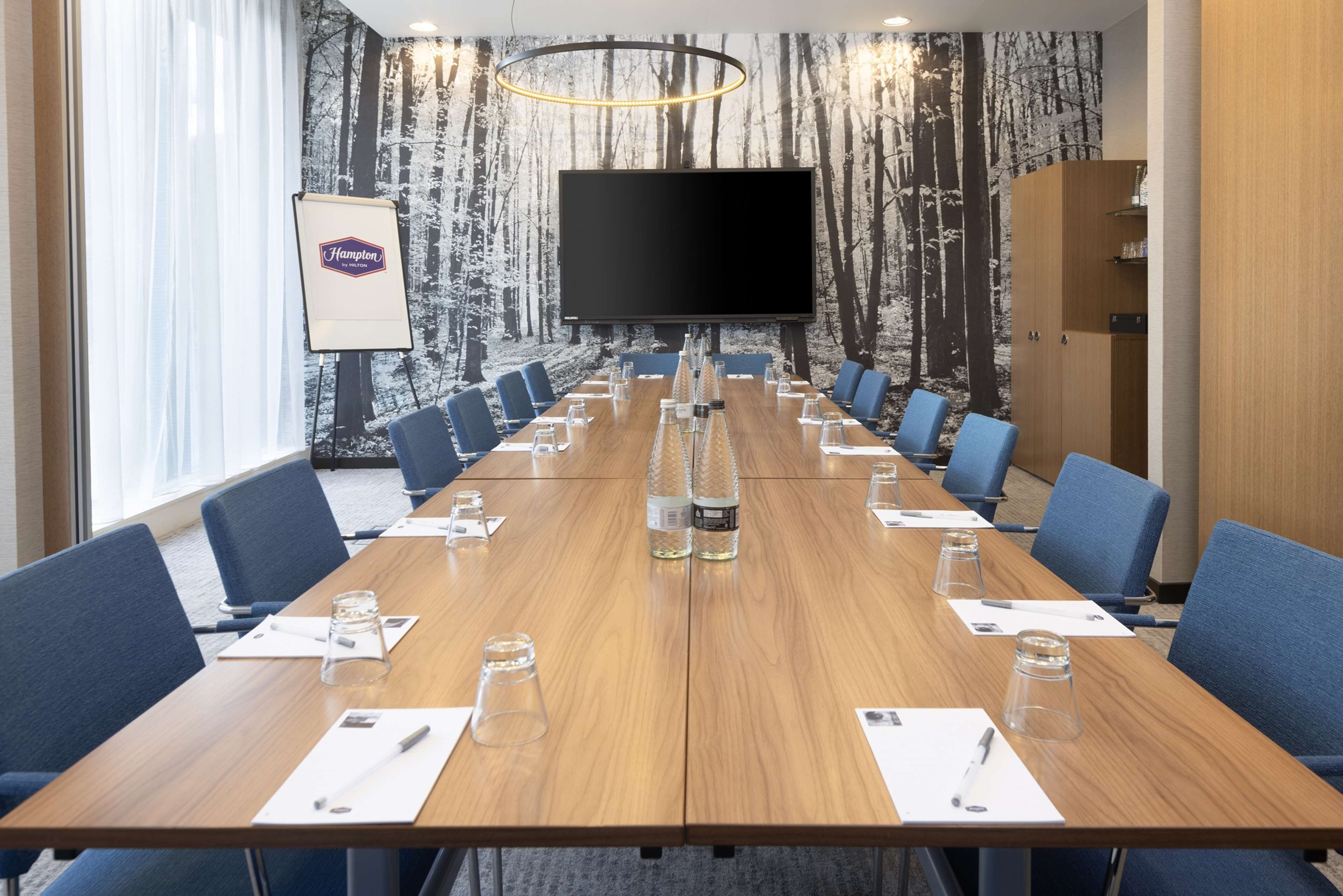 A conference room with a long table, blue chairs, a large screen, and a forest-themed wall.