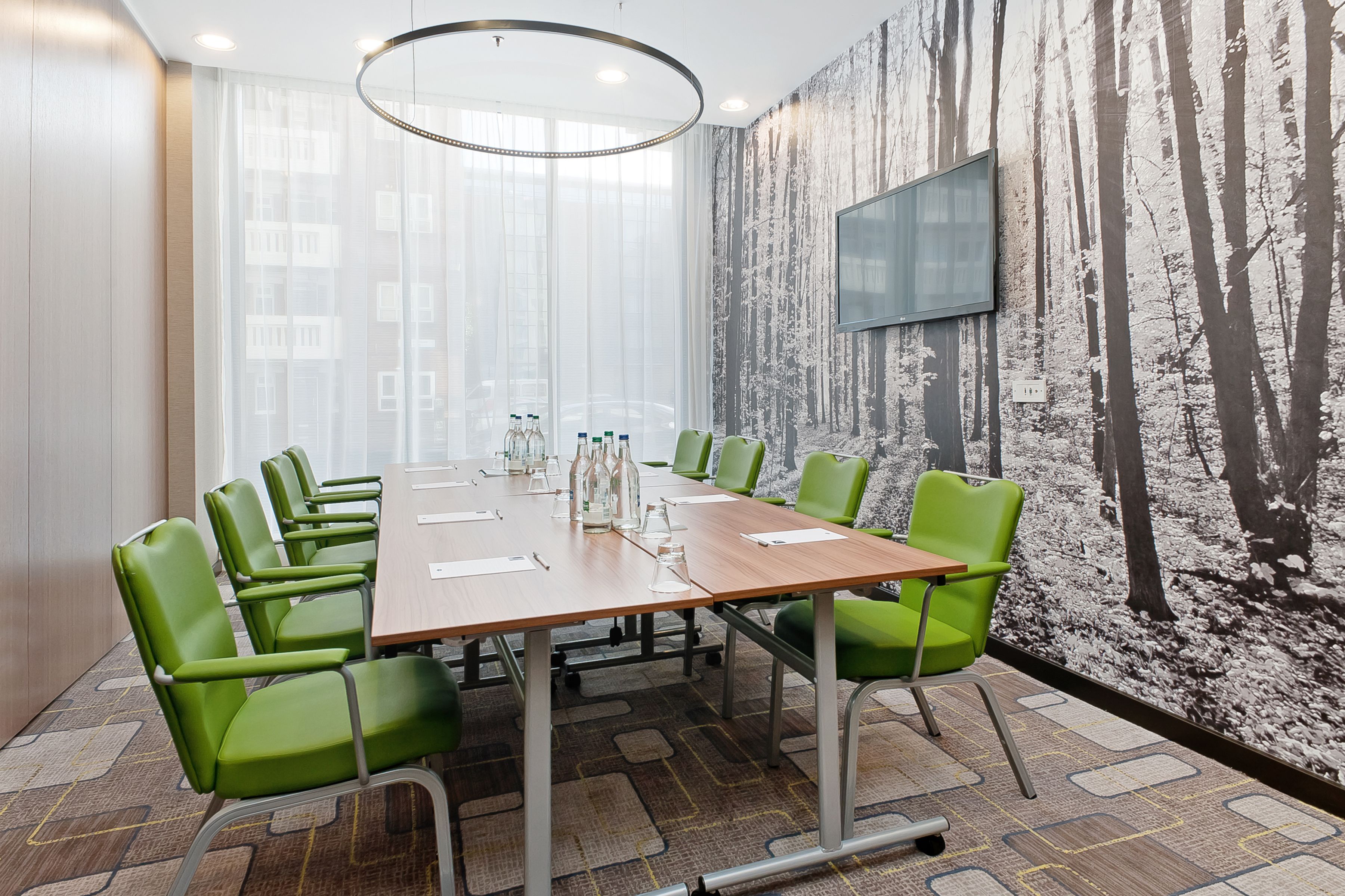 A conference room with a large table, green chairs, a mounted TV, and a forest-themed wall.