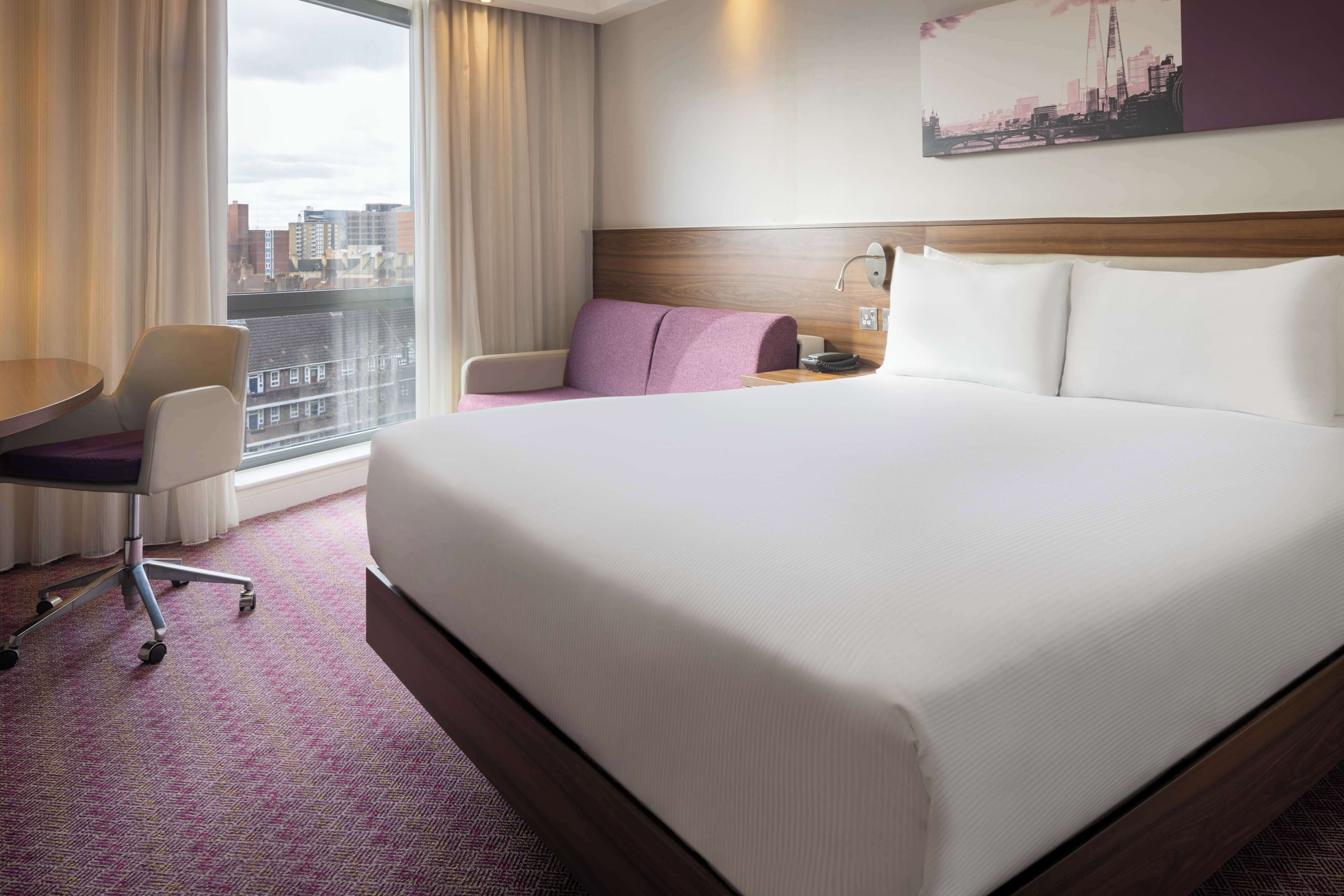A hotel room with a large bed, a desk with a chair, a window with curtains, and a cityscape view.