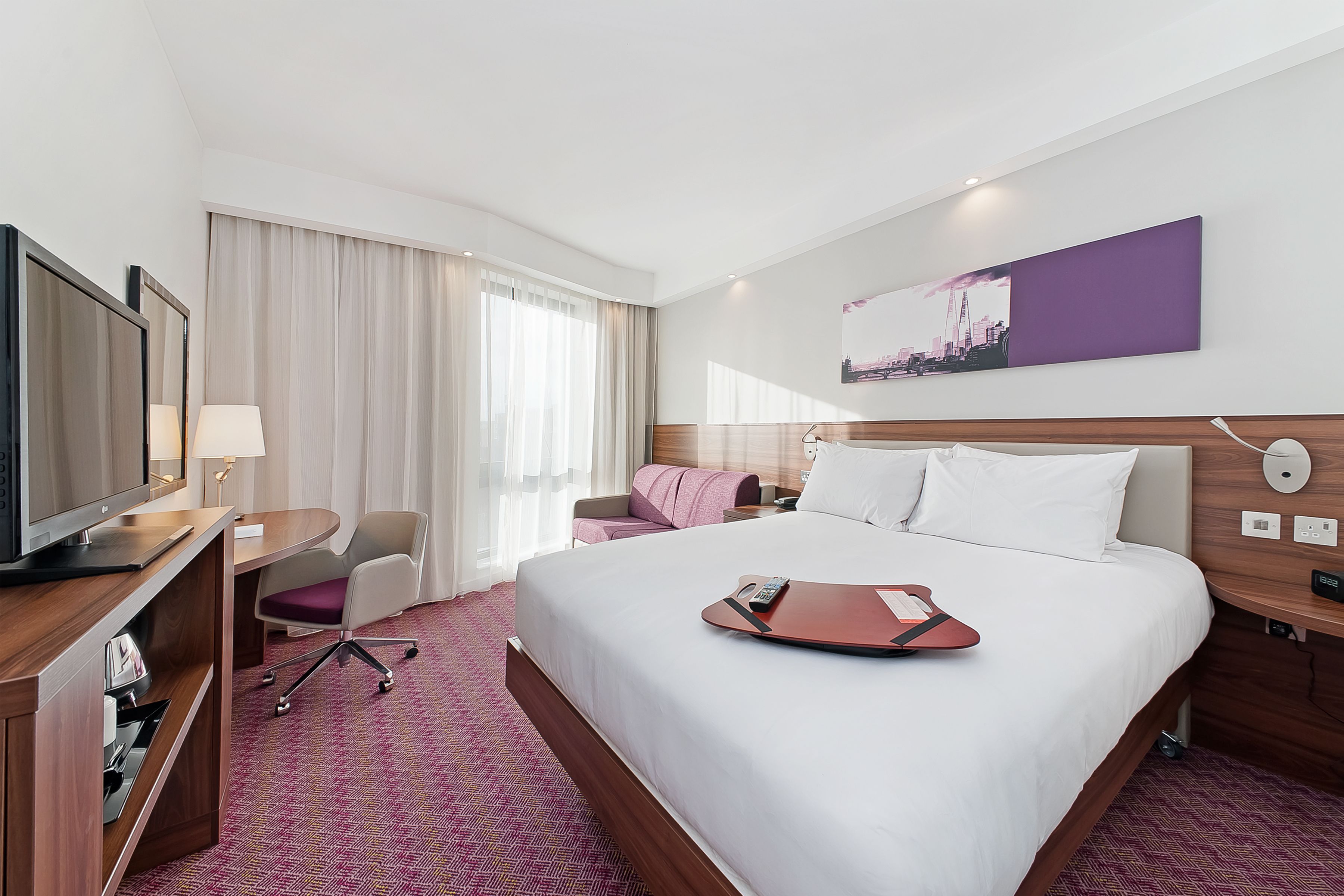 A hotel room with a large bed, a desk with a television, a chair, and a window with curtains.