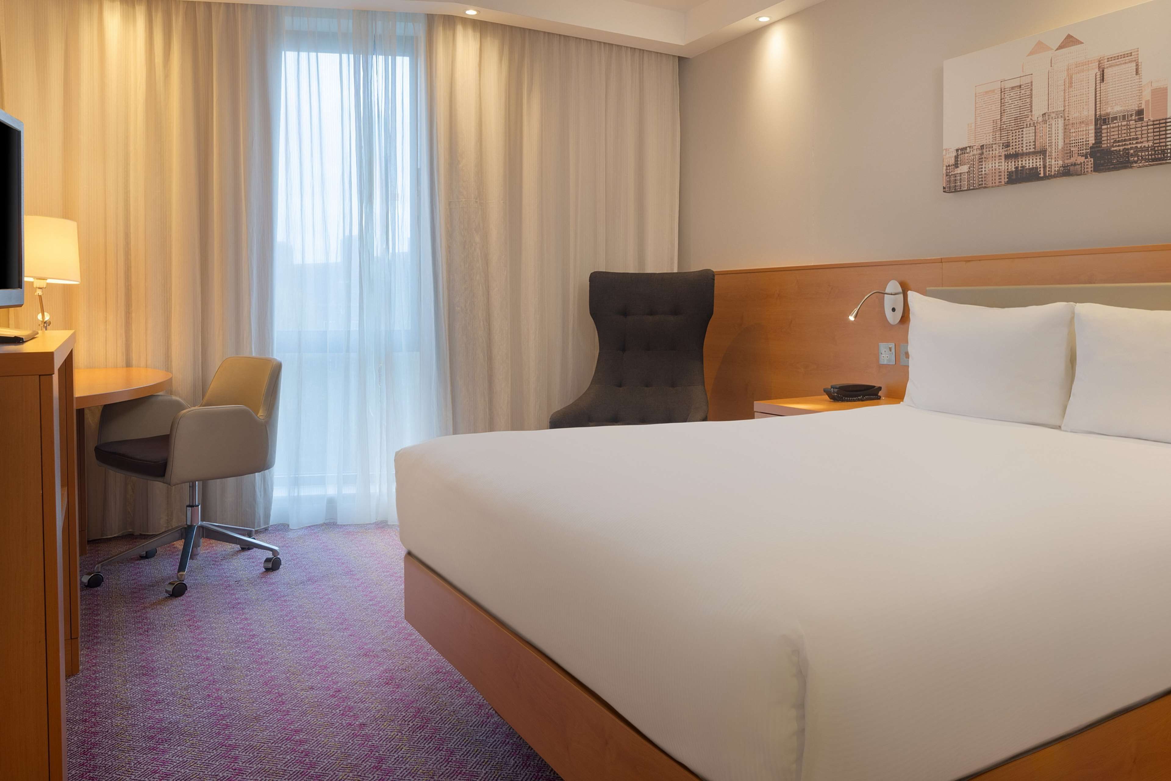 Une chambre d’hôtel avec un grand lit, un bureau avec une chaise, une télévision et une œuvre d’art représentant un paysage urbain accrochée au mur.