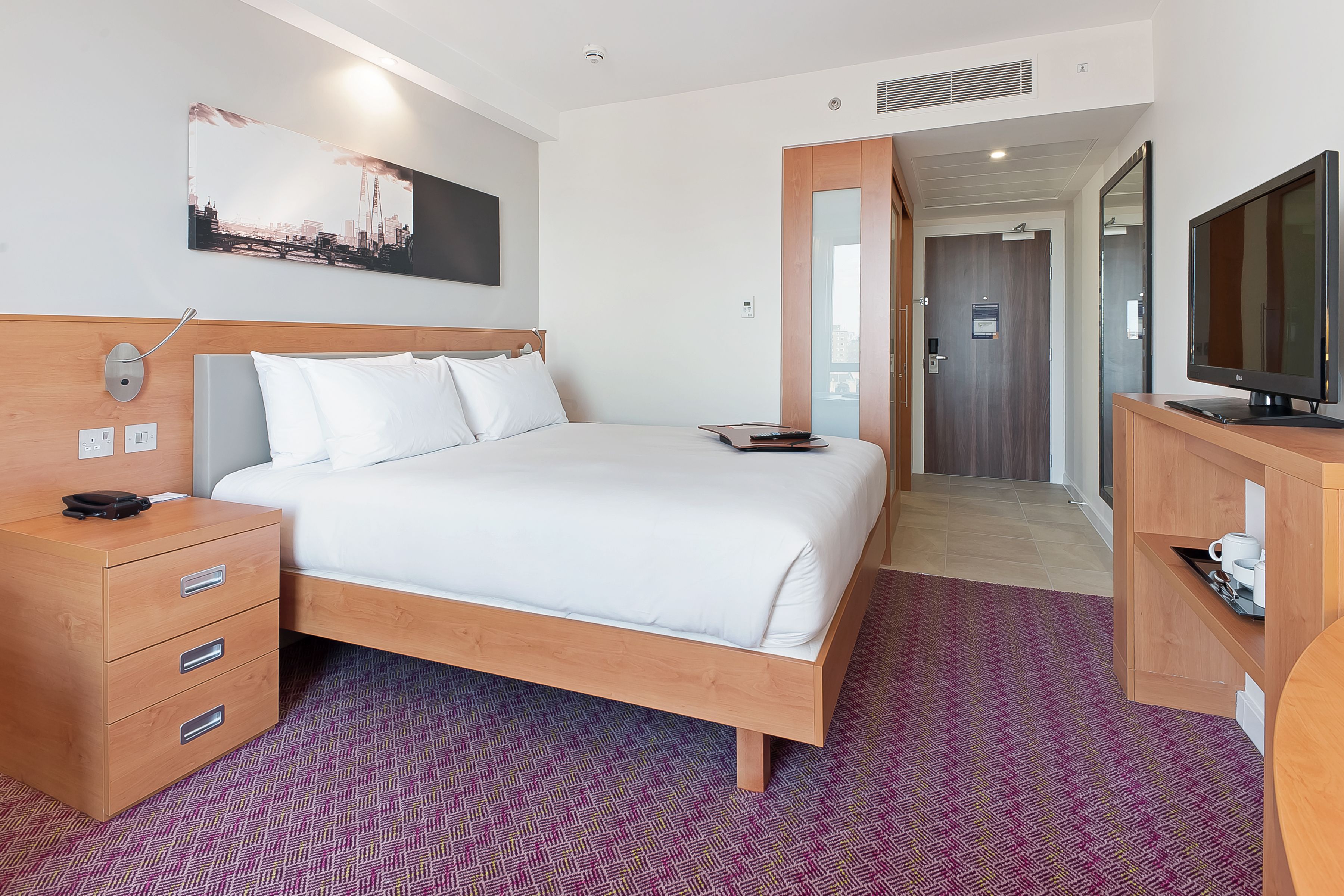 A hotel room with a bed, a wooden nightstand, a TV, and a cityscape painting on the wall.