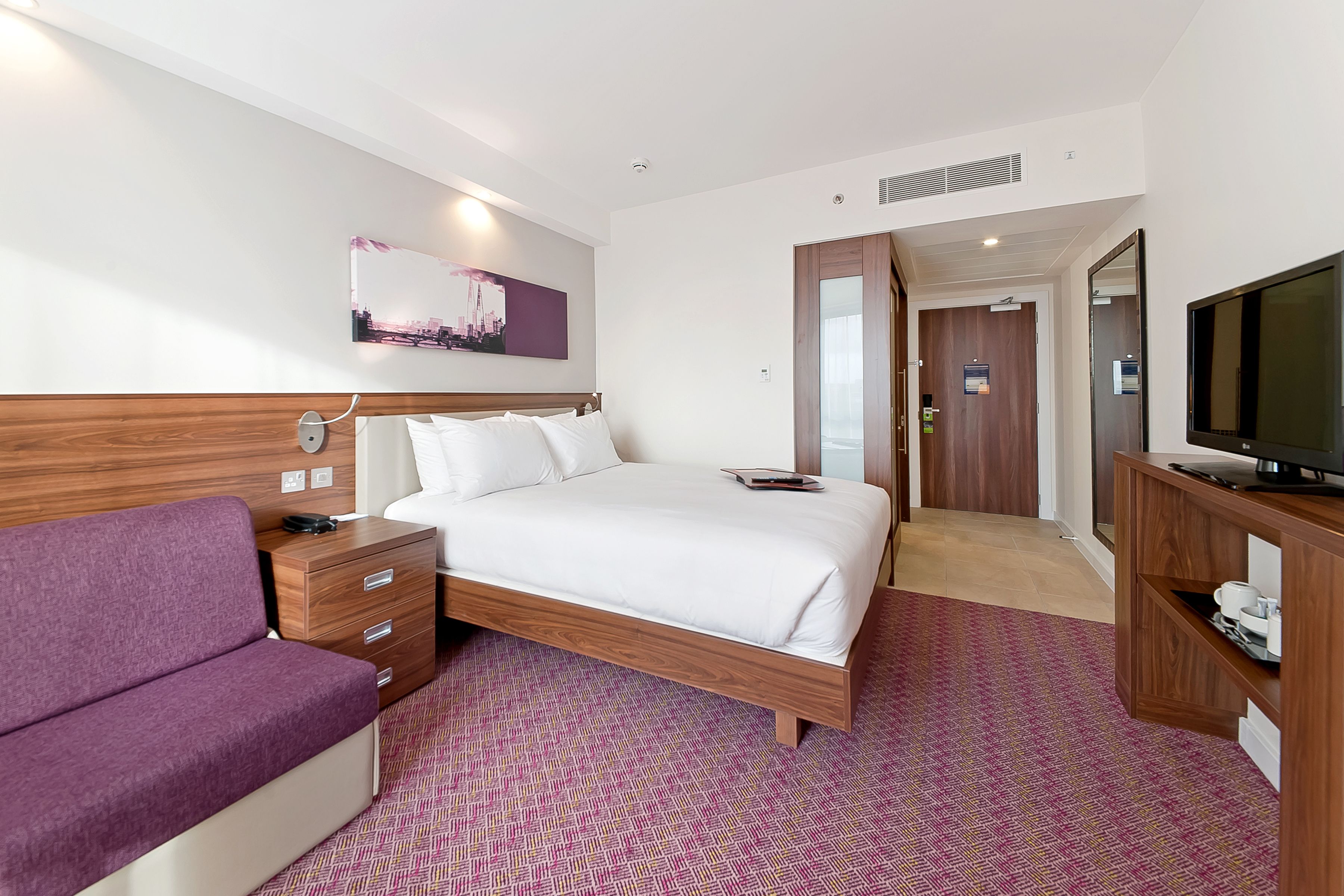 Una habitación de hotel con una cama, un sofá morado, un televisor y una pintura de un paisaje urbano en la pared.