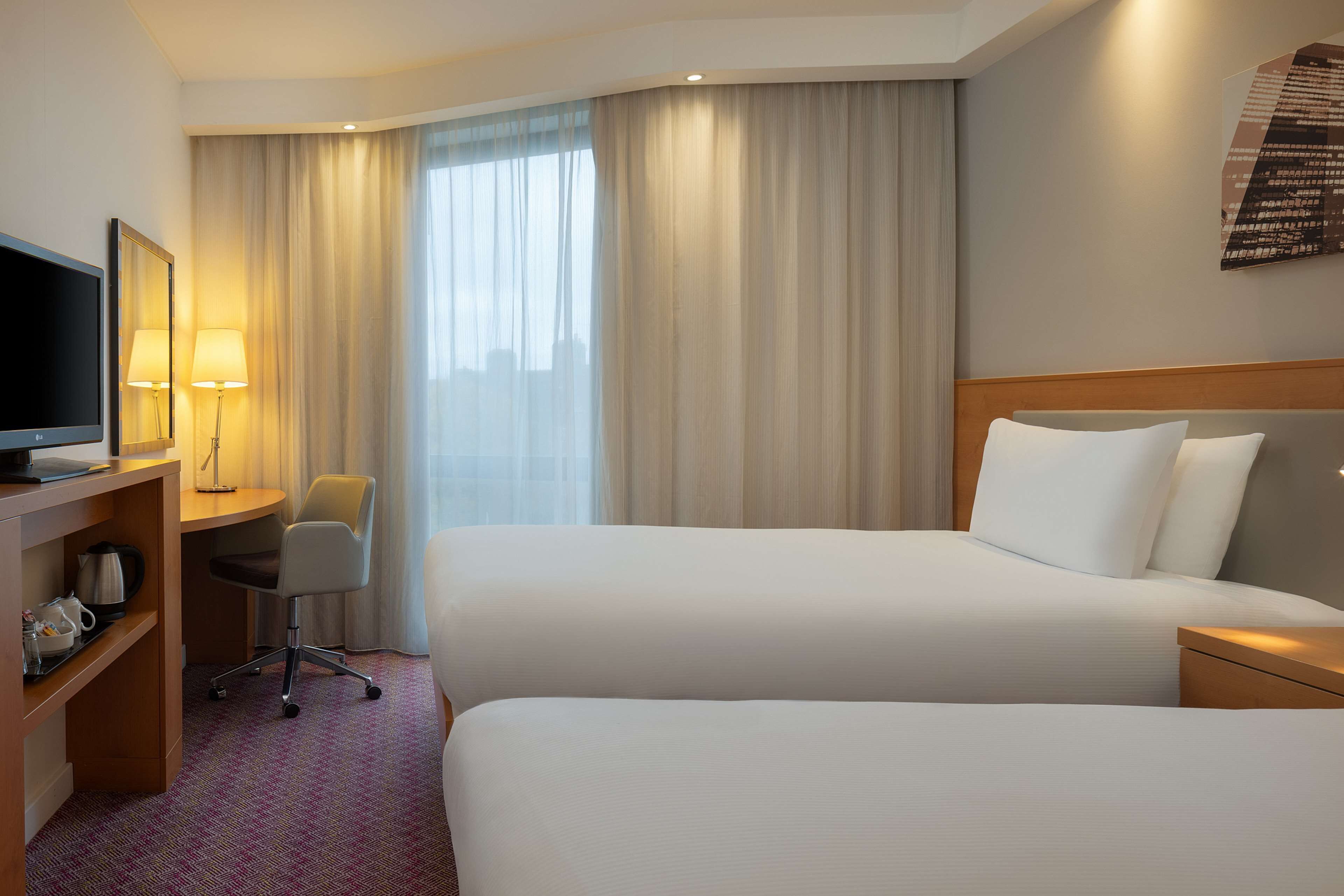 A hotel room with two beds, a desk with a chair, a television, and a large window with curtains.