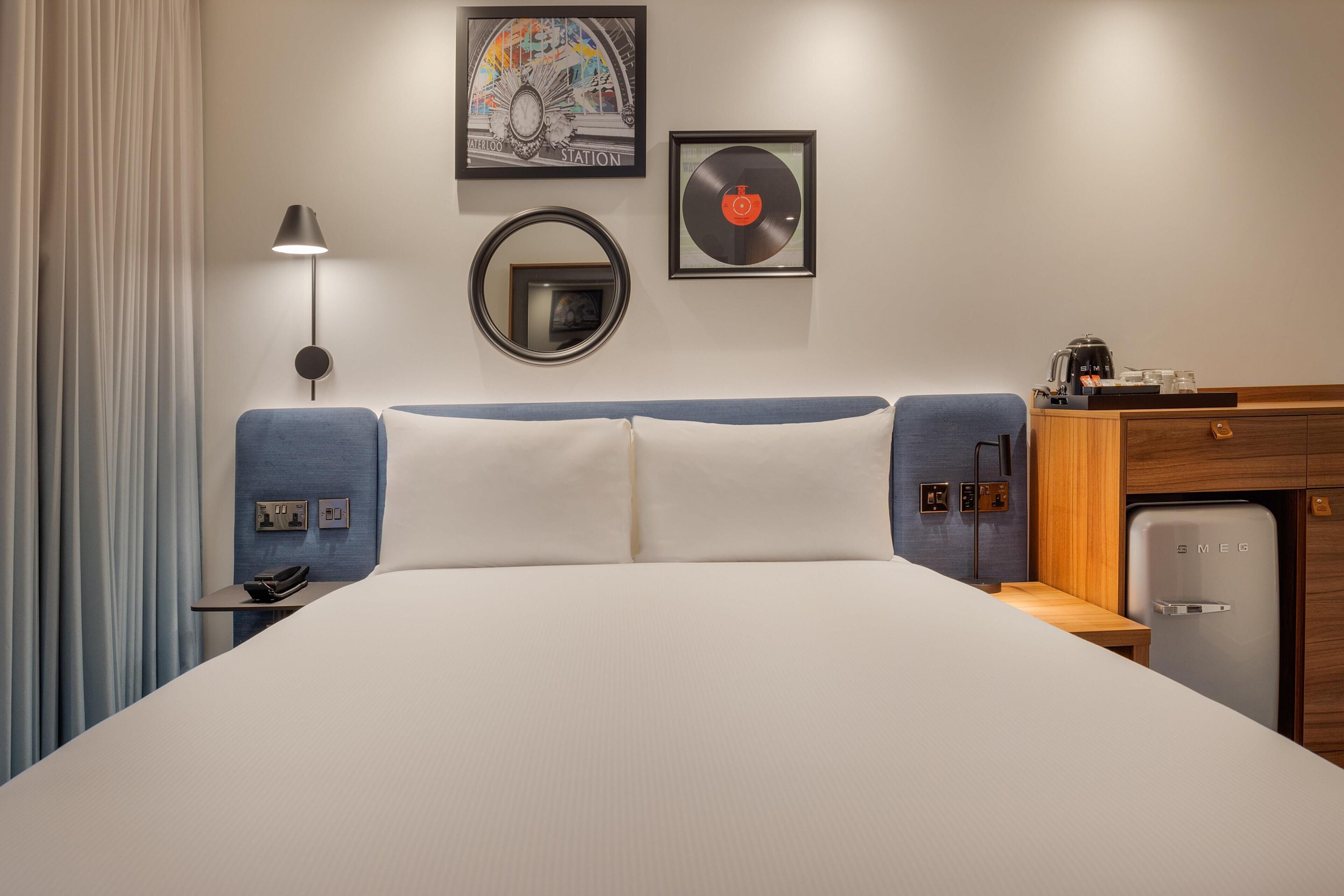 A hotel room with a bed, a desk with a lamp, a refrigerator, and wall decorations including a clock and a vinyl record.