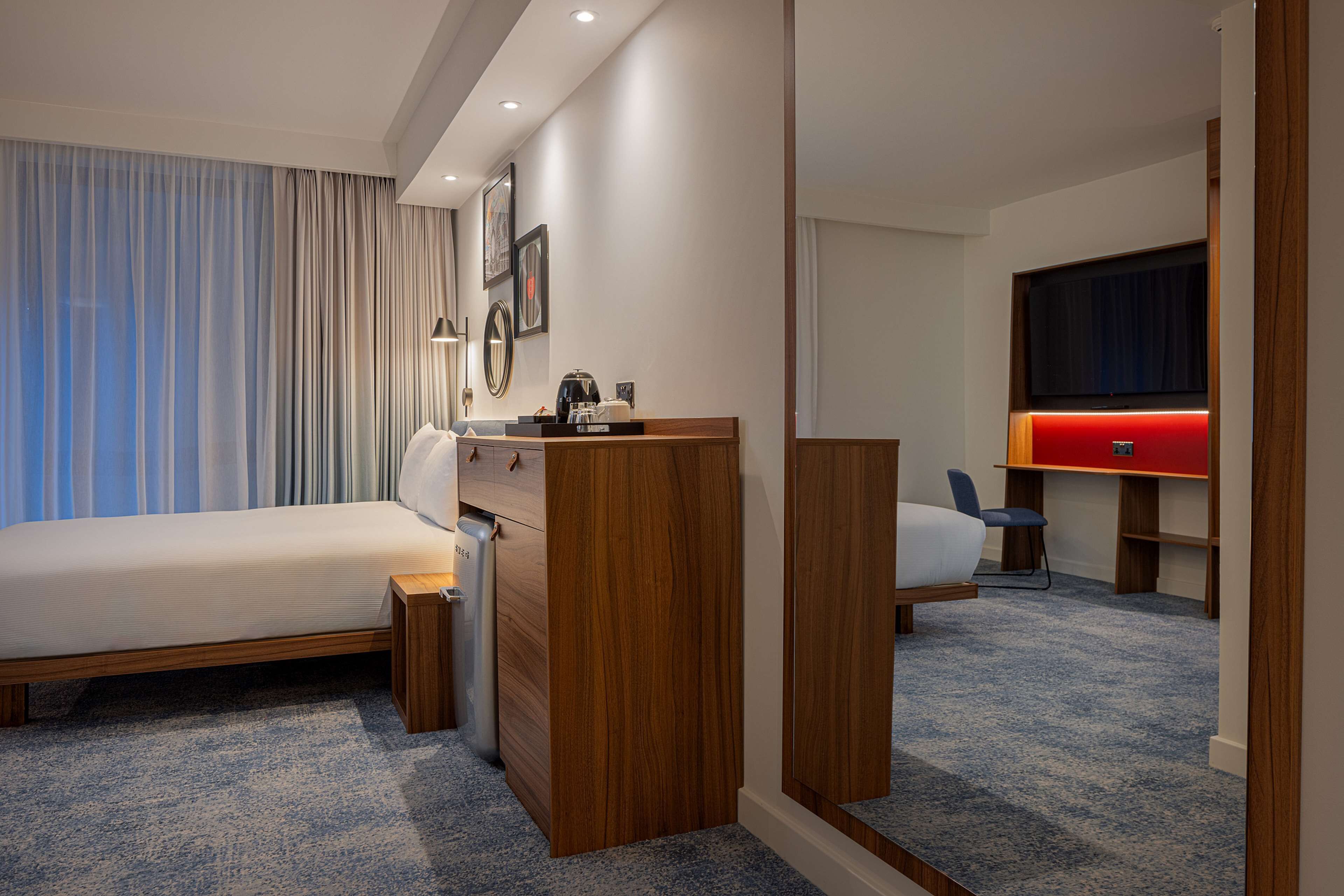 A hotel room with a bed, a desk, a chair, a TV, and a red accent wall.