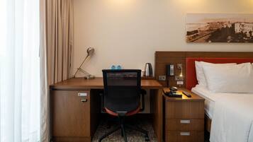 Une chambre d’hôtel avec un bureau en bois, une chaise de bureau noire, un lit avec une literie blanche, une table de chevet avec une lampe, une télécommande et un tableau encadré au mur.