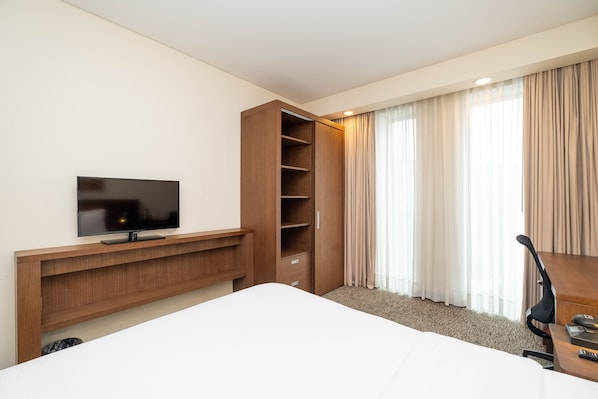 Habitación de hotel con una cama, un televisor sobre un soporte de madera, un escritorio con una silla y un ventanal con cortinas.