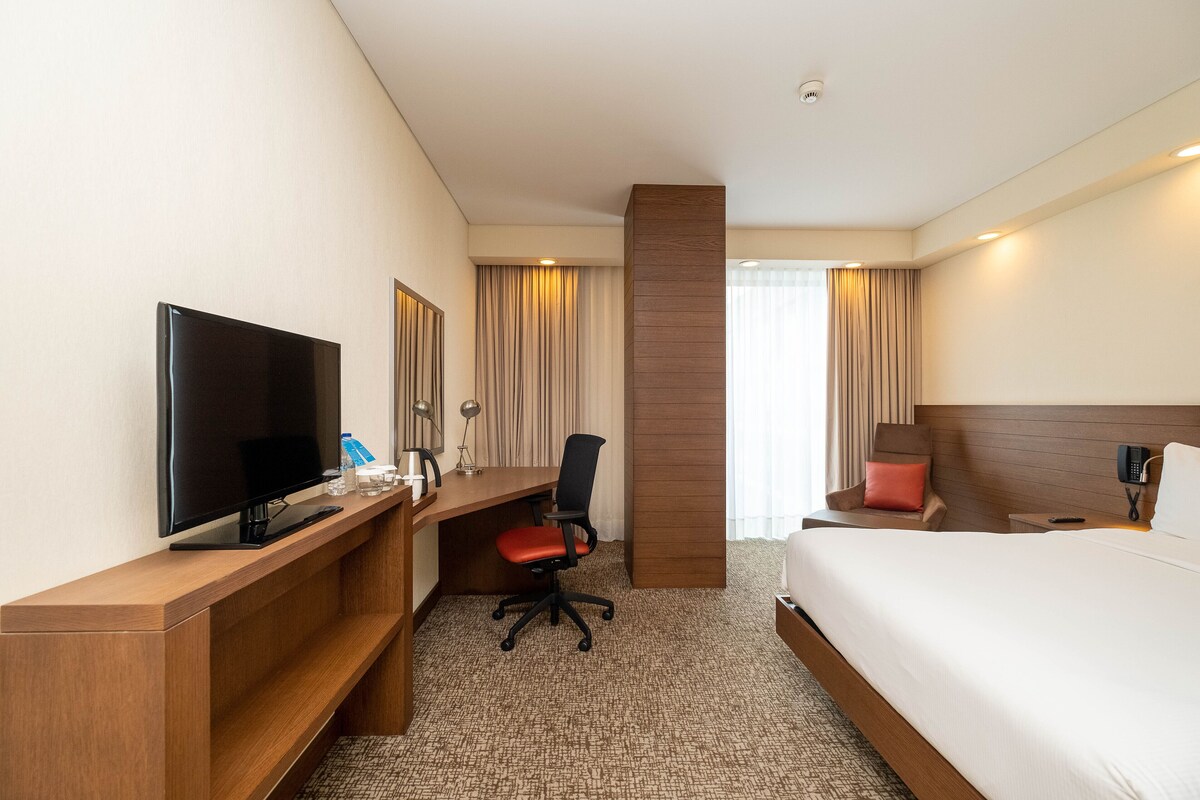 Una habitación de hotel con una cama, un escritorio con televisor, una silla y un ventanal con cortinas.