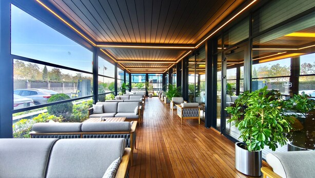 Un balcón interior moderno con suelo de madera, asientos tapizados en gris y ventanas amplias con vistas al estacionamiento y a la vegetación.