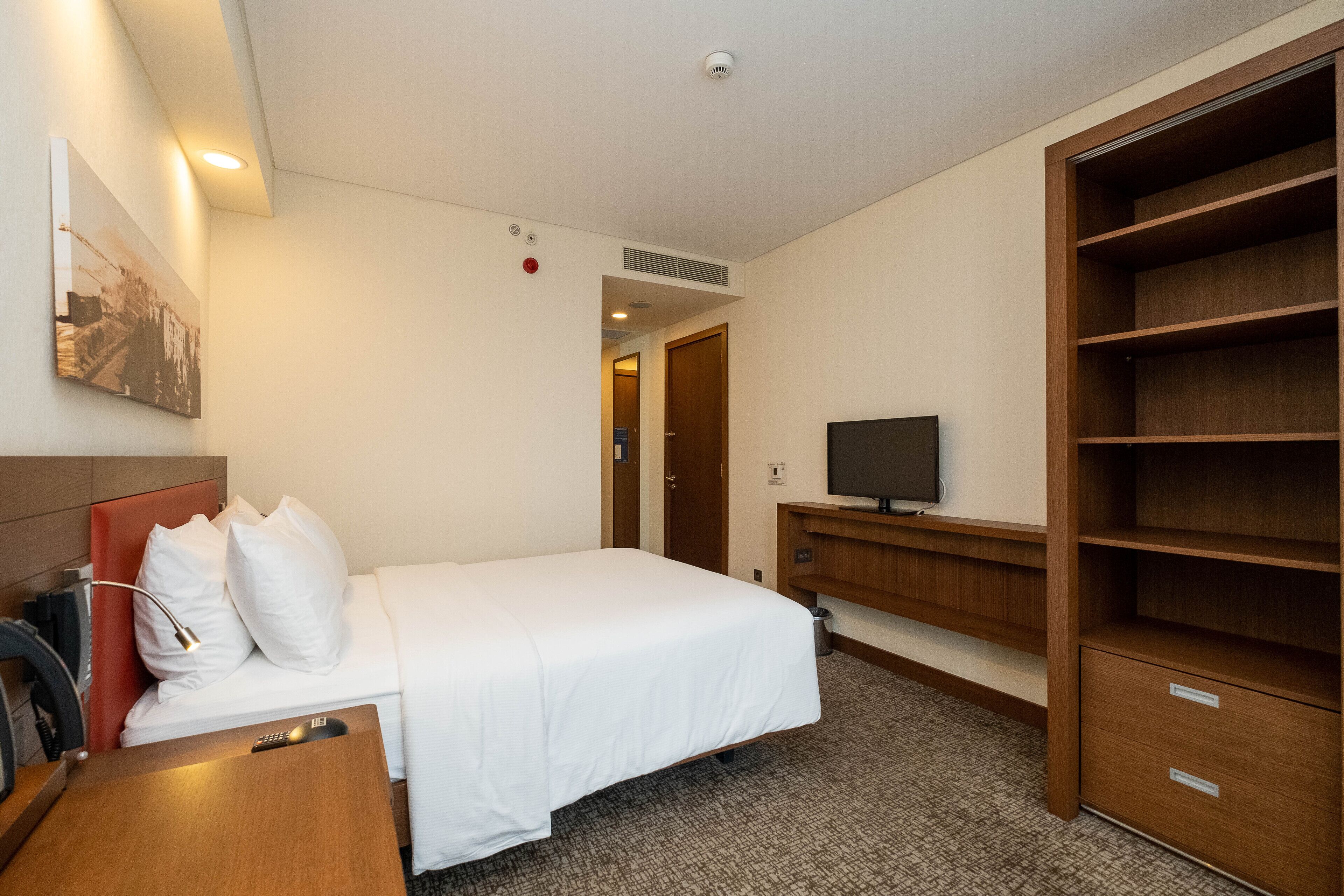 A hotel room with a bed, a desk with a lamp, a TV, a wardrobe, and a painting on the wall.