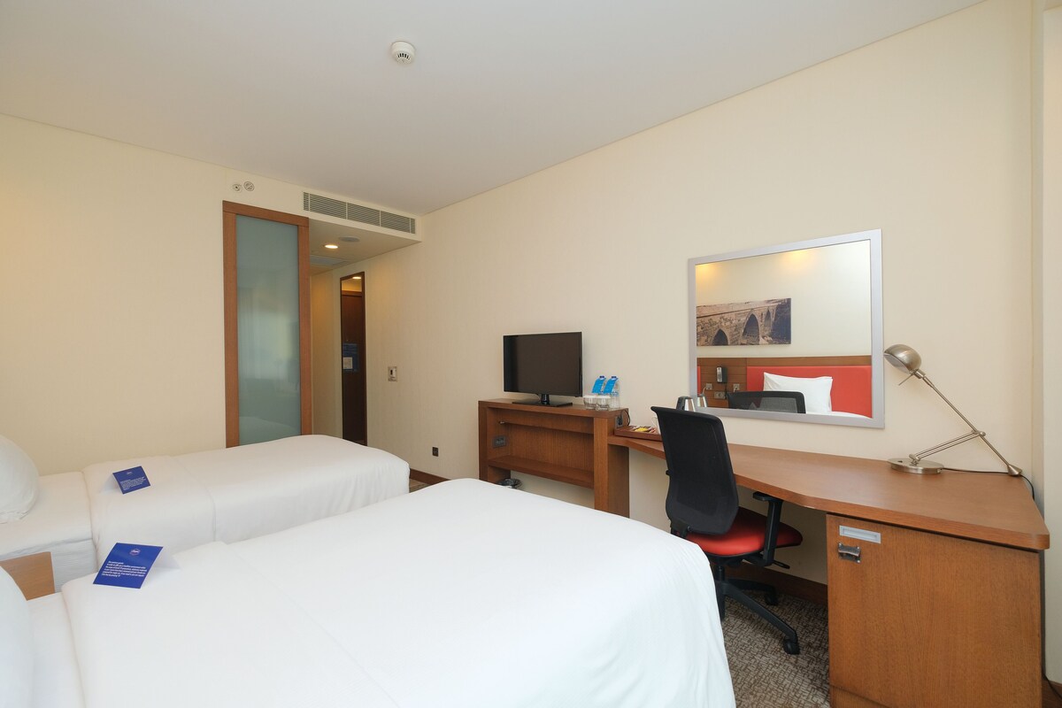 Habitación de hotel con dos camas individuales, un escritorio con una computadora, una silla, una lámpara de escritorio y un espejo.