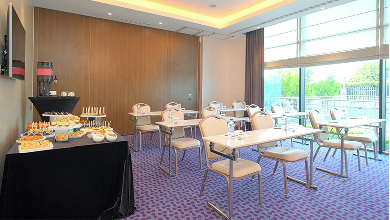 Sala de conferência com mesas e cadeiras, mesa de buffet com comida e janelas amplas com vista.