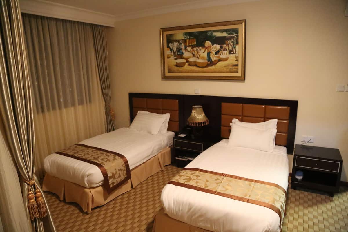 A hotel room with two beds, a nightstand, a lamp, a painting on the wall, and a check-in counter.