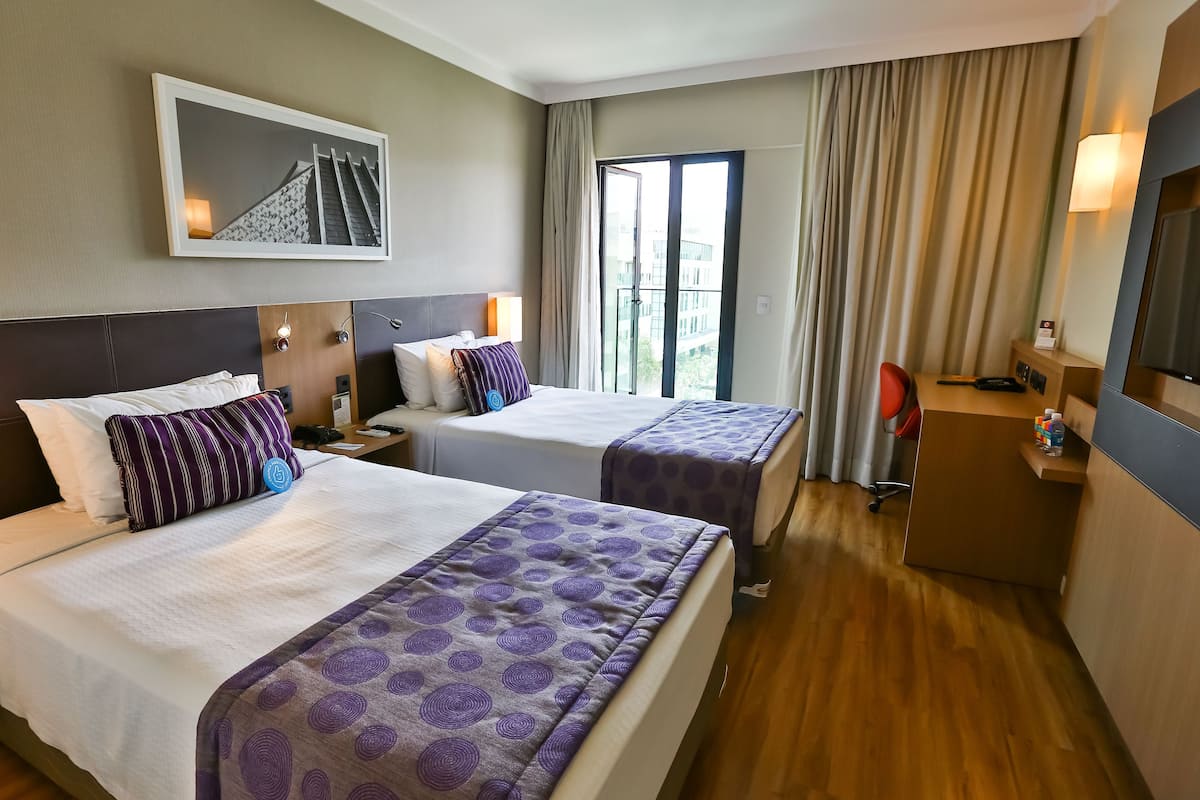 A hotel room with two beds, a TV, a desk, and a large window with curtains.