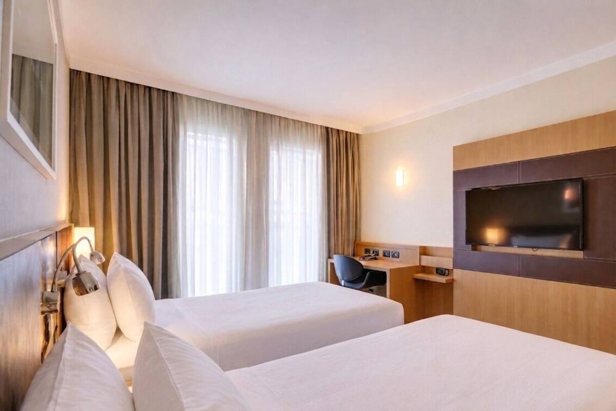 A hotel room with two beds, a desk, a chair, a TV, and a window with curtains.