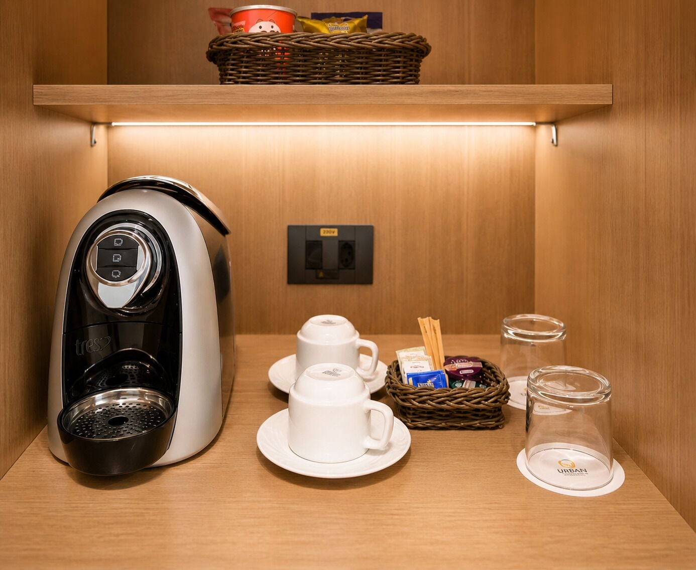 A kitchenette with a coffee maker, two white cups, a basket of snacks, and a glass jar.