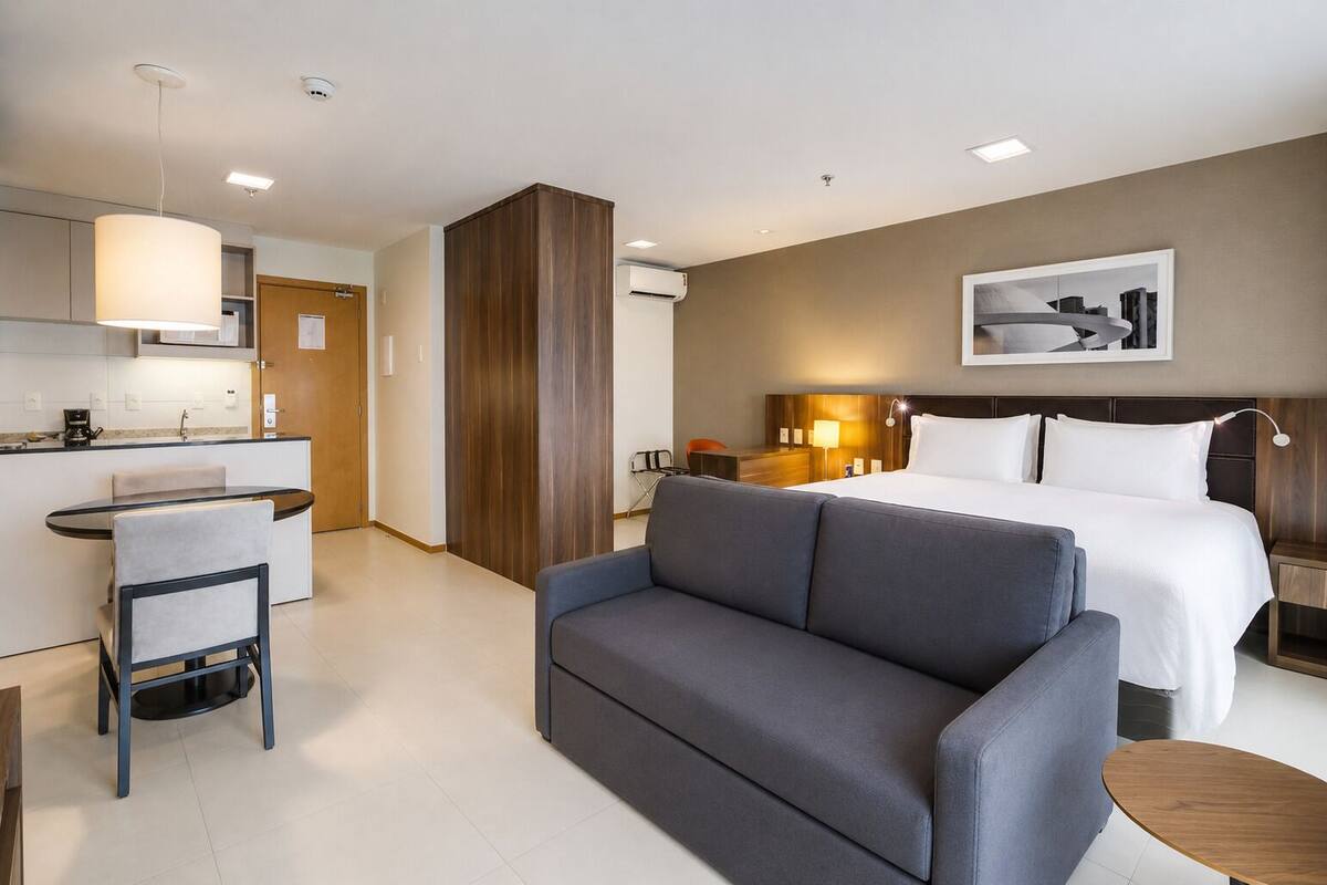 A hotel room with a bed, a sofa, a desk with a chair, a small table, and a wall-mounted picture.