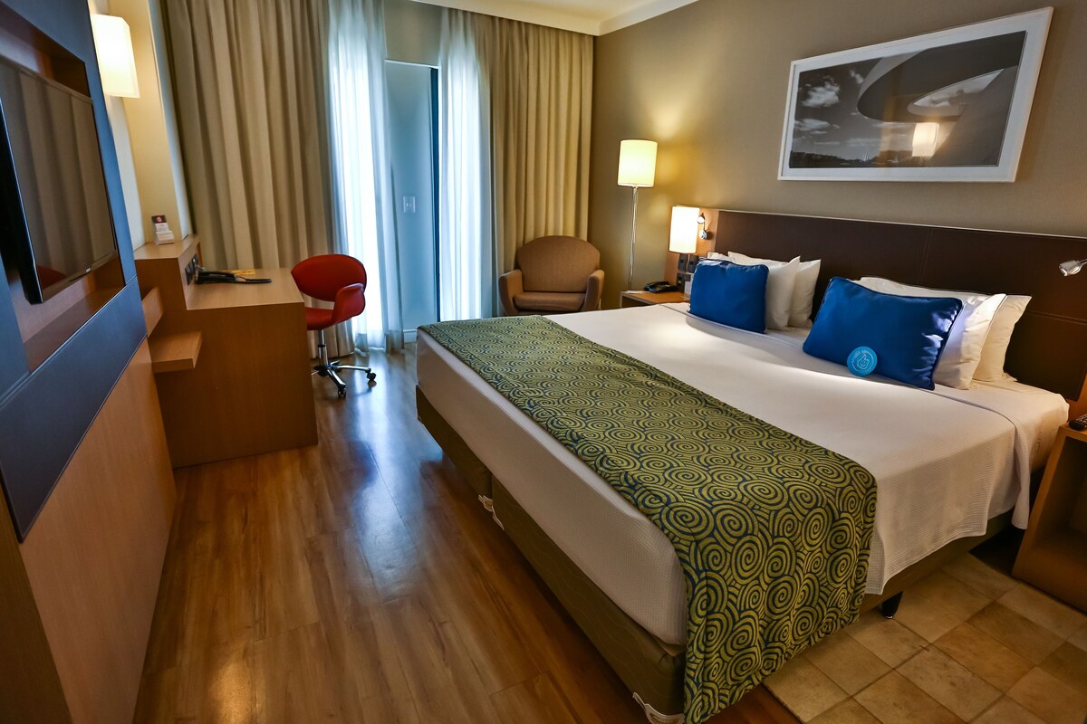 Habitación de hotel con cama, escritorio, silla, lámpara y televisor.