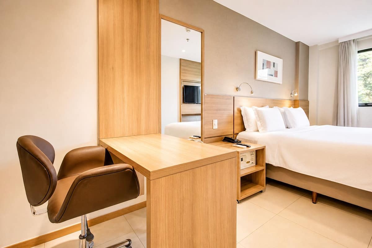 A hotel room with a wooden desk, two leather chairs, a bed with white linens, a large mirror, and a framed picture on the wall.