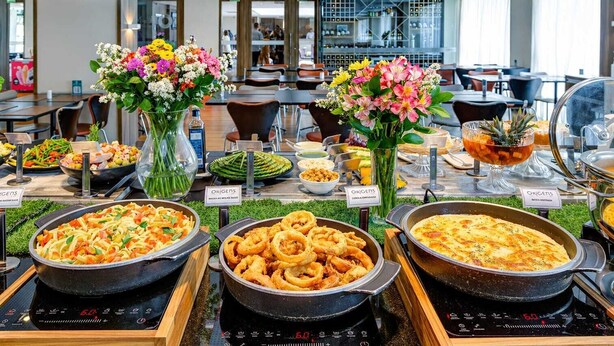 A buffet with various food options, including pasta, fried items, and a baked dish, in a modern dining area with tables and chairs.