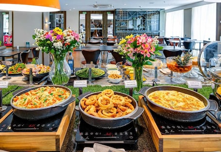 A buffet with various food options, including pasta, fried items, and a baked dish, in a modern dining area with tables and chairs.
