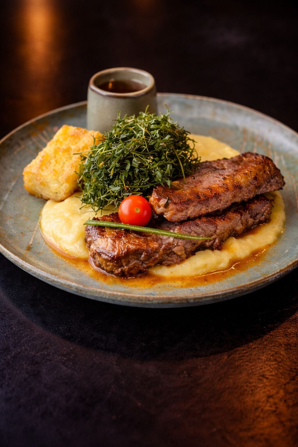Een bord met steak, gepureerde aardappelen, een kleine tomaat en een takje kruiden.