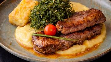 Een bord met steak, gepureerde aardappelen, een kleine tomaat en een takje kruiden.