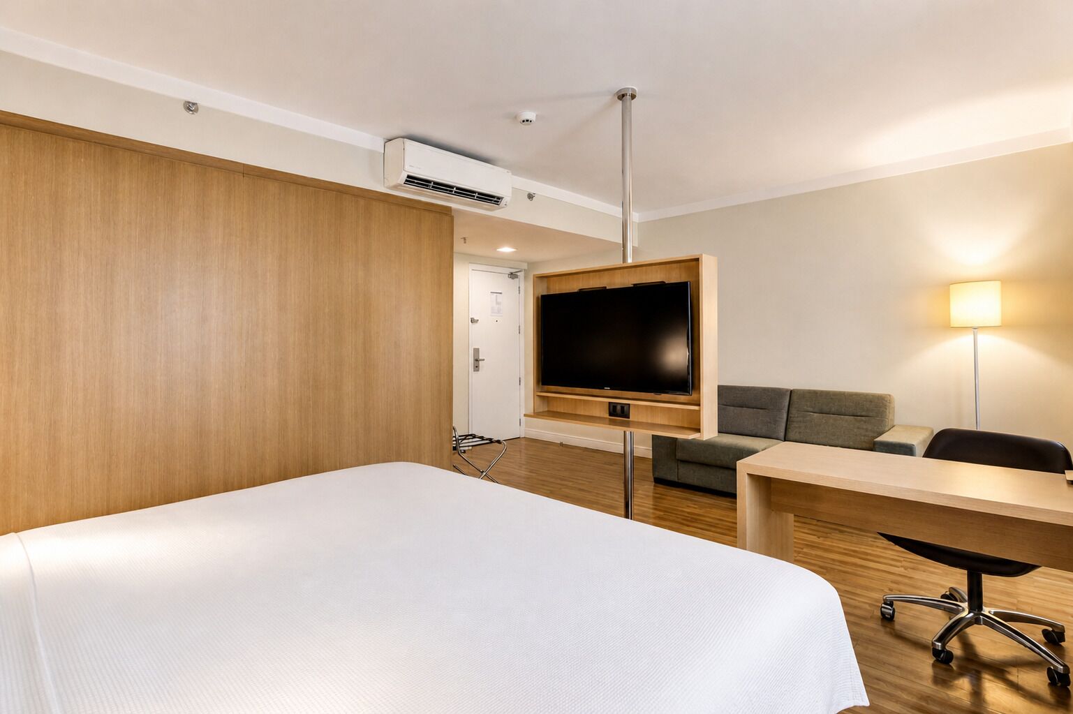 A hotel room with a bed, a TV mounted on the wall, a desk, a chair, a sofa, and a lamp.