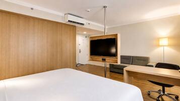 A hotel room with a bed, a TV mounted on the wall, a desk, a chair, a sofa, and a lamp.