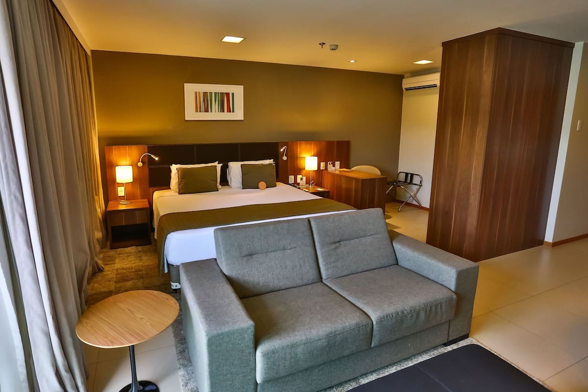 A hotel room with a bed, a sofa, a desk, a chair, a lamp, and a wall-mounted artwork.